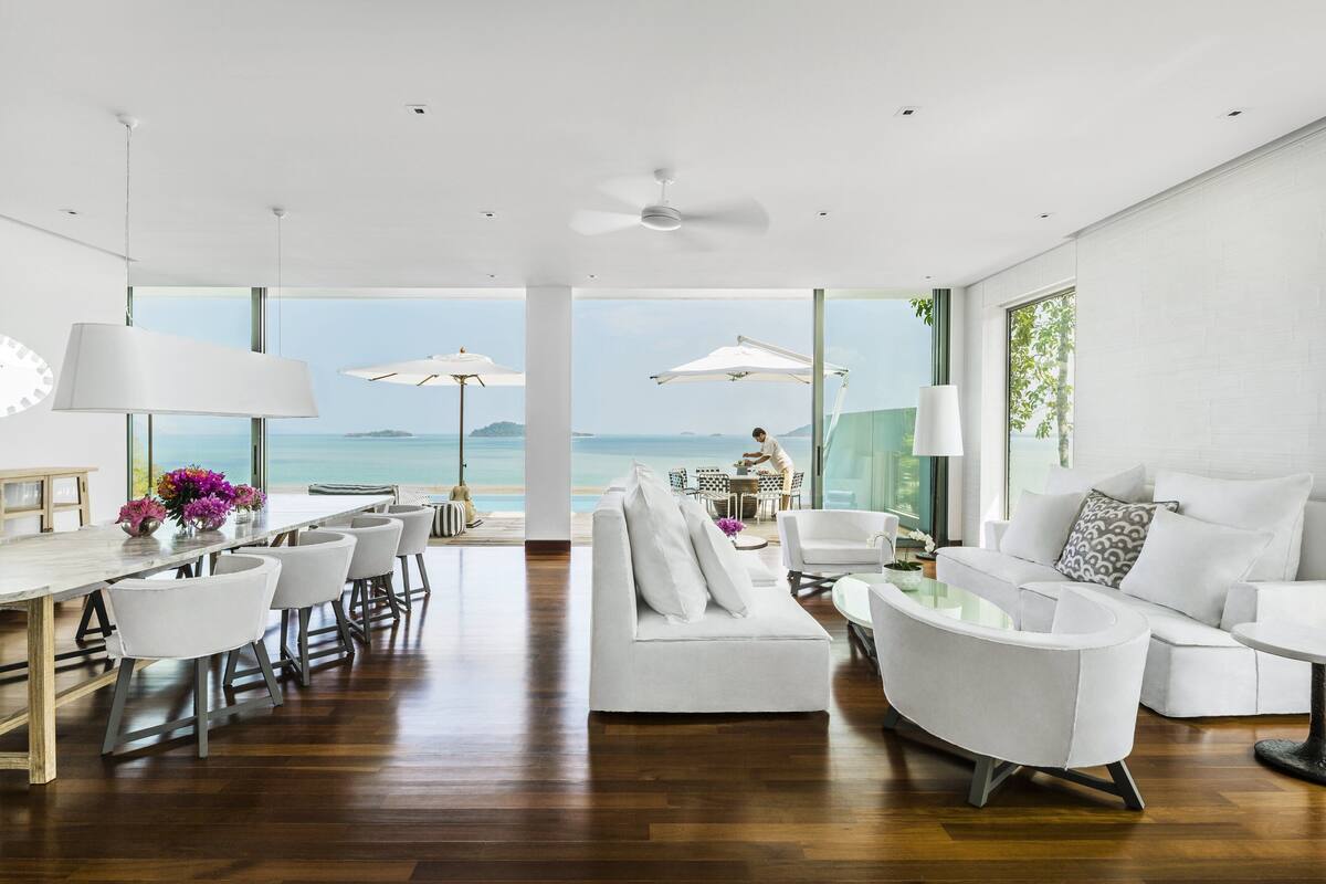 Een moderne woonkamer met een groot raam met uitzicht op zee, een witte bank, een eethoek met stoelen en een plafondventilator.