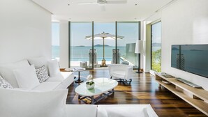 A modern living room with a large TV, white furniture, a glass coffee table, and a view of the sea.
