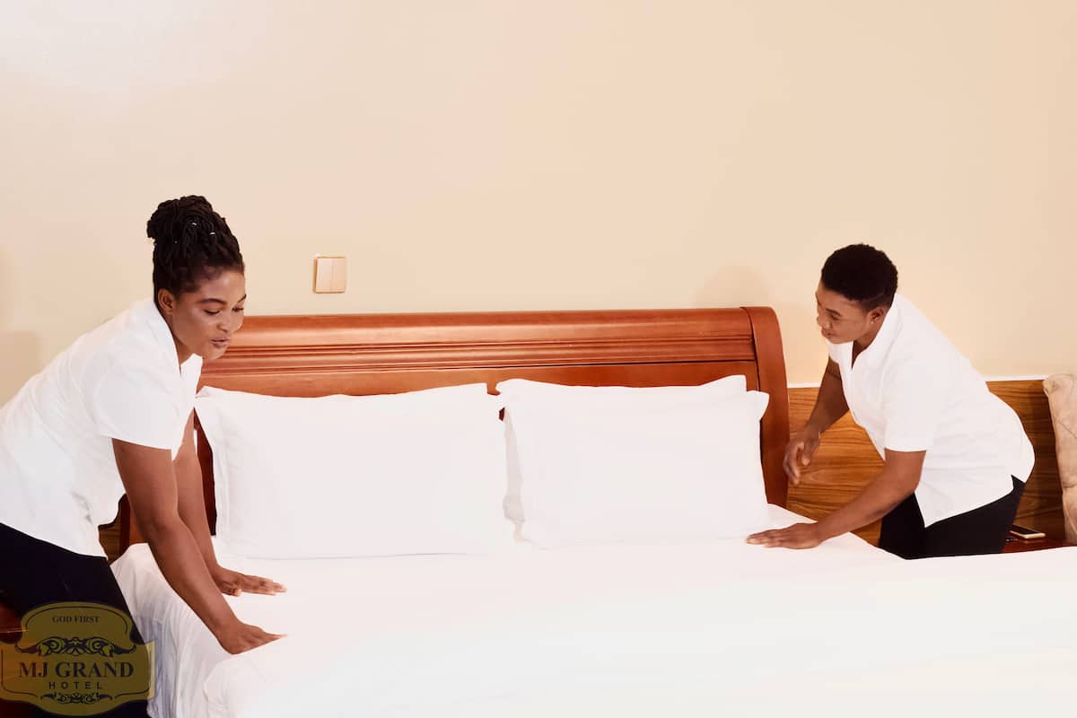 Two individuals arranging a bed in a hotel room with a wooden headboard and white bedding.