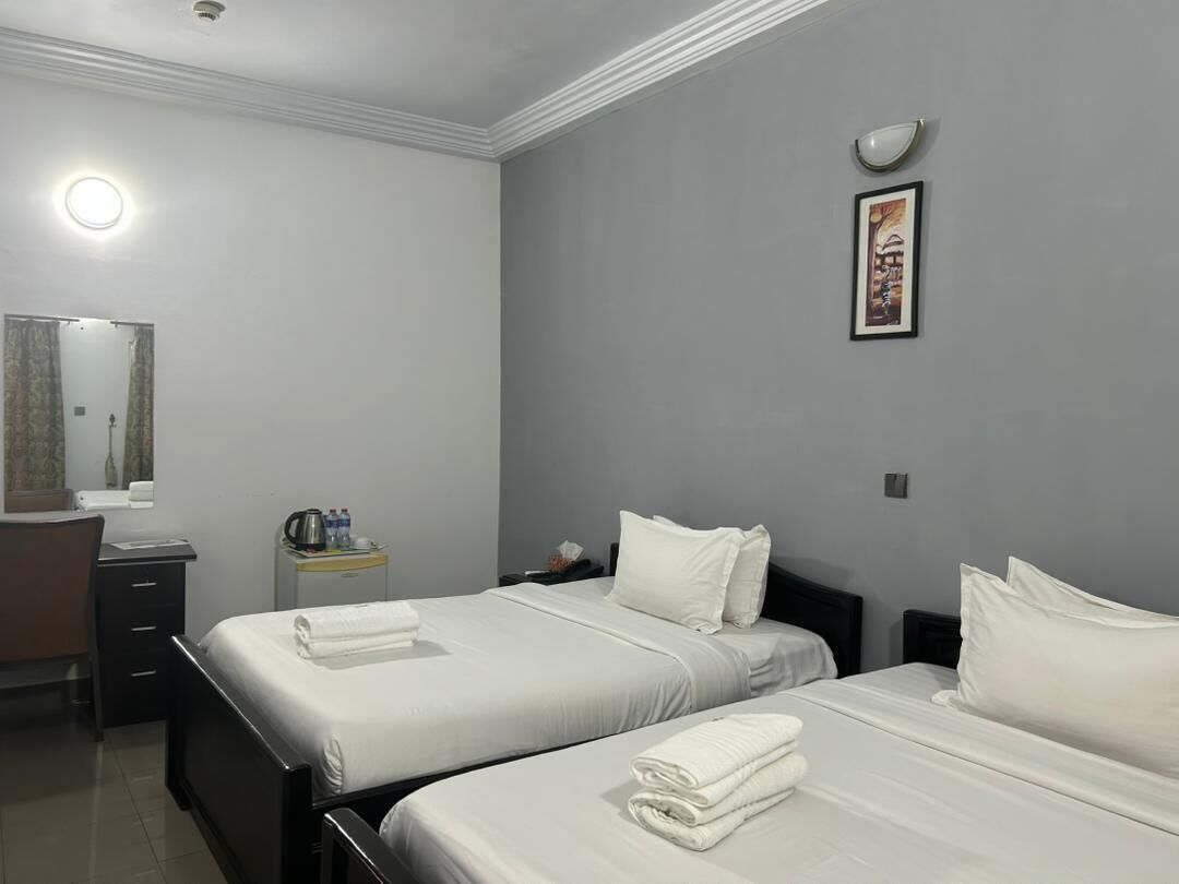A hotel room with two beds, a desk, a chair, a small table with a kettle, and a framed picture on the wall.