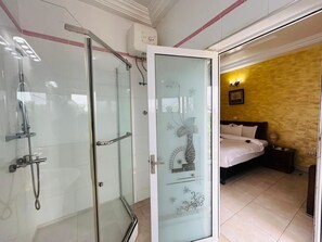 A bathroom with a glass shower enclosure, a bed with a white bedspread, and a wall with a decorative design. - MJ Grand Hotel (Accra)