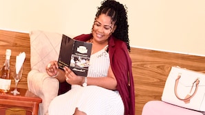 A person seated on a cushioned chair, holding a magazine, with a wooden floor and a small table with a bottle and glass. - MJ Grand Hotel (Accra)