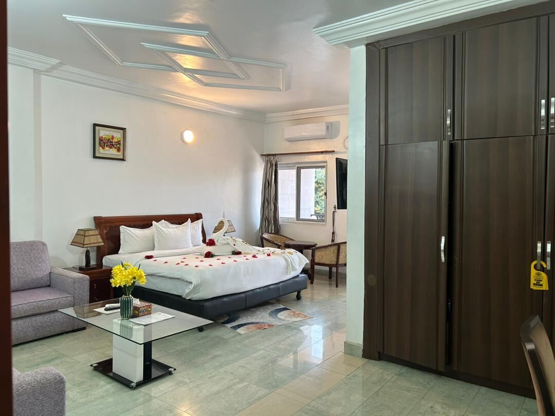 A hotel room with a bed, a sofa, a coffee table with flowers, a wardrobe, and a window with curtains.