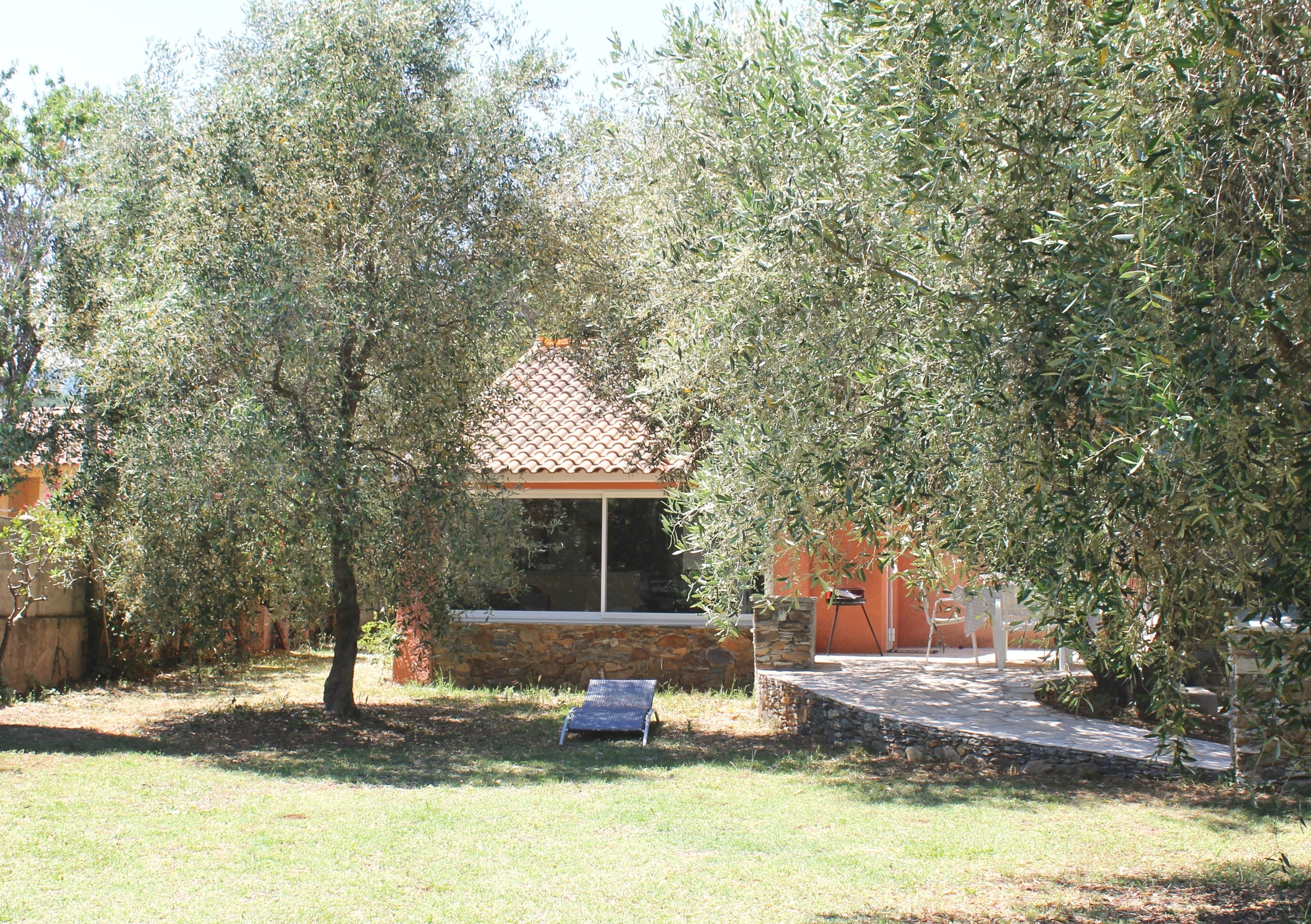 Une maison avec un toit en tuiles, entourée d’oliviers, et un petit patio avec une chaise et une table.