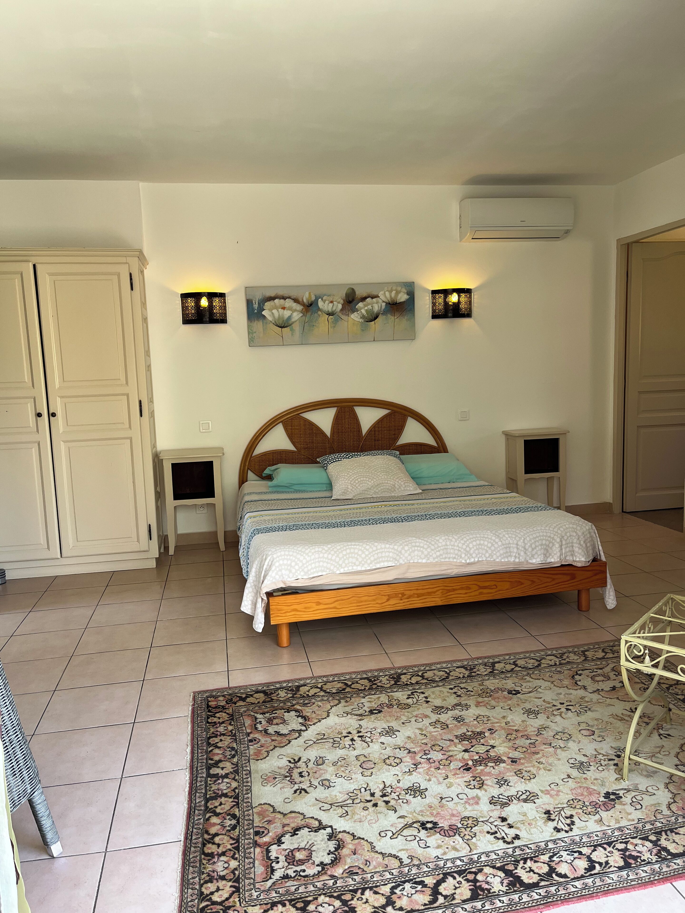 A bedroom with a wooden bed, two bedside tables, a wall-mounted air conditioner, and a large floral rug.