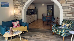 A living room with a sofa, a coffee table, and a patterned armchair. A kitchen is visible through an archway. - Résidence Le Clos des Oliviers (Sorbo-Ocagnano)