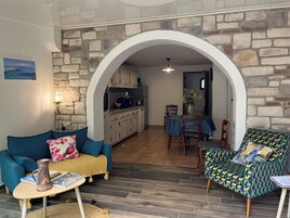 Sala de estar com sofá, mesa de centro e poltrona estampada. Cozinha visível por um arco.