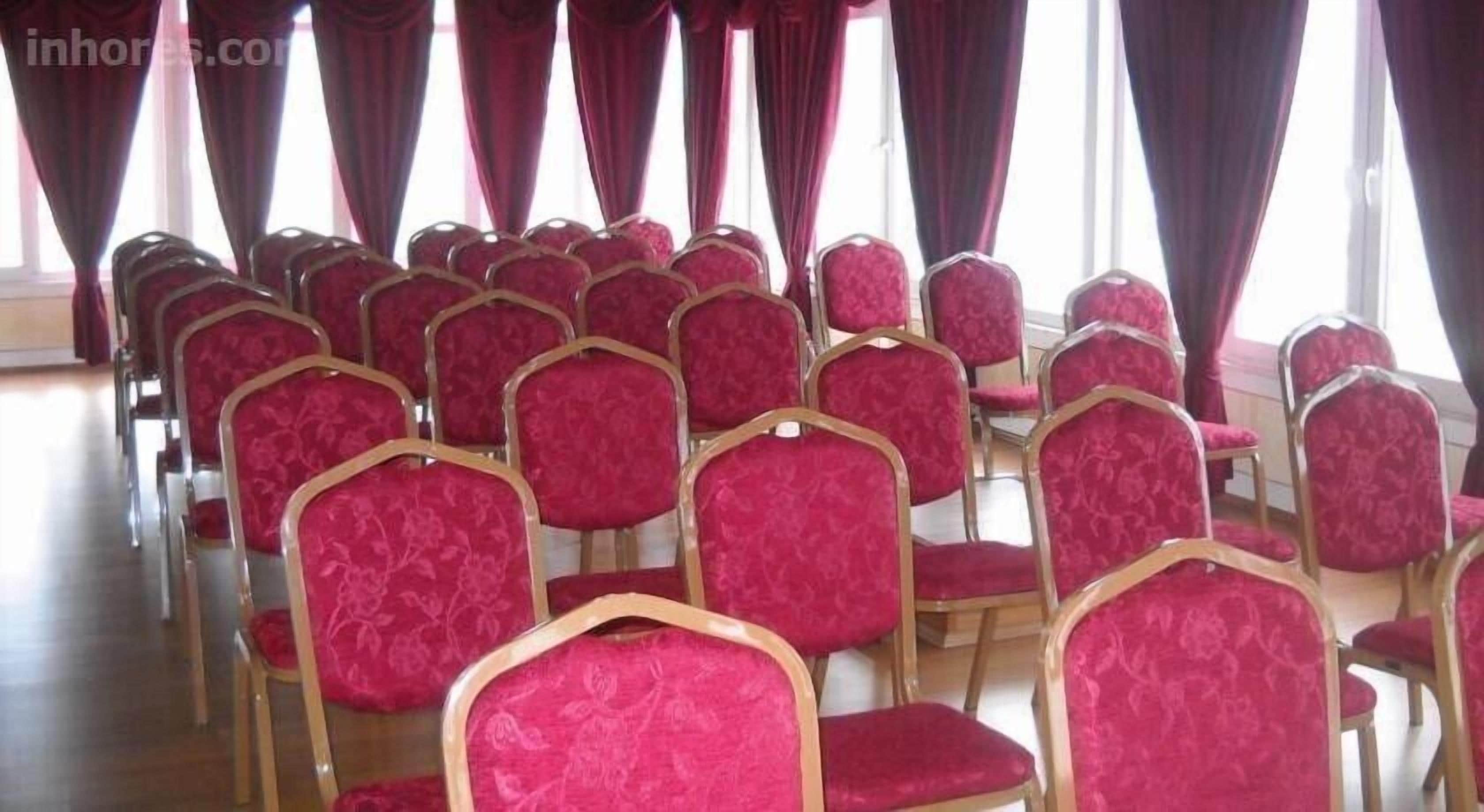 Rows of red upholstered chairs with gold frames, arranged in a room with large windows and red curtains.