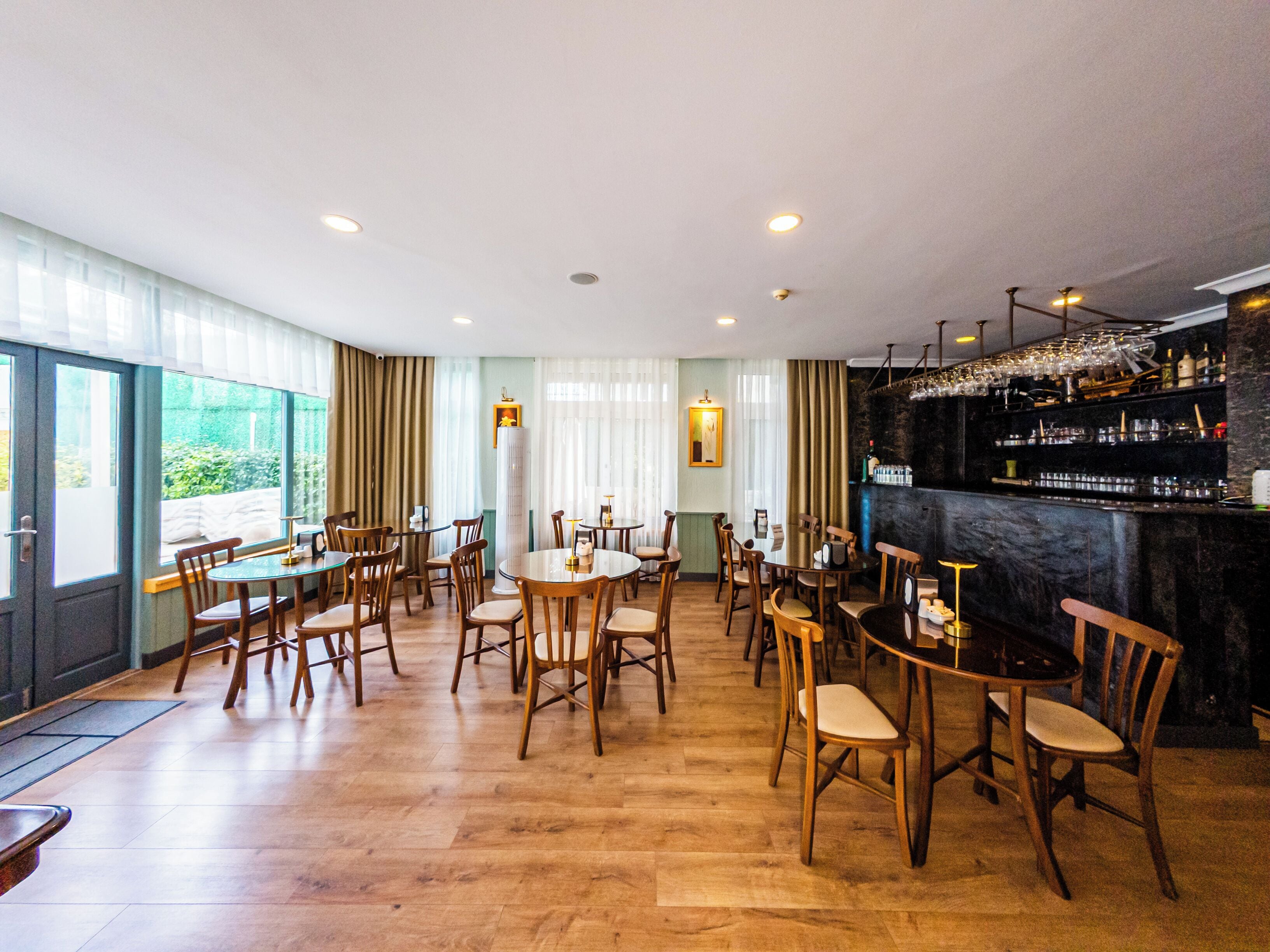 A restaurant with multiple tables and chairs, a bar area with shelves of glasses and bottles, and large windows with curtains.