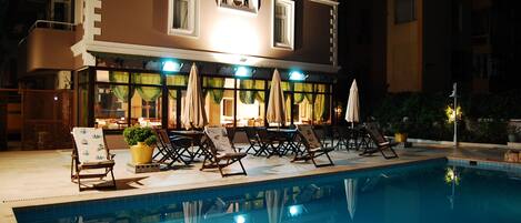 A hotel with a pool area featuring lounge chairs and umbrellas, a well-lit exterior, and a reflection in the pool.