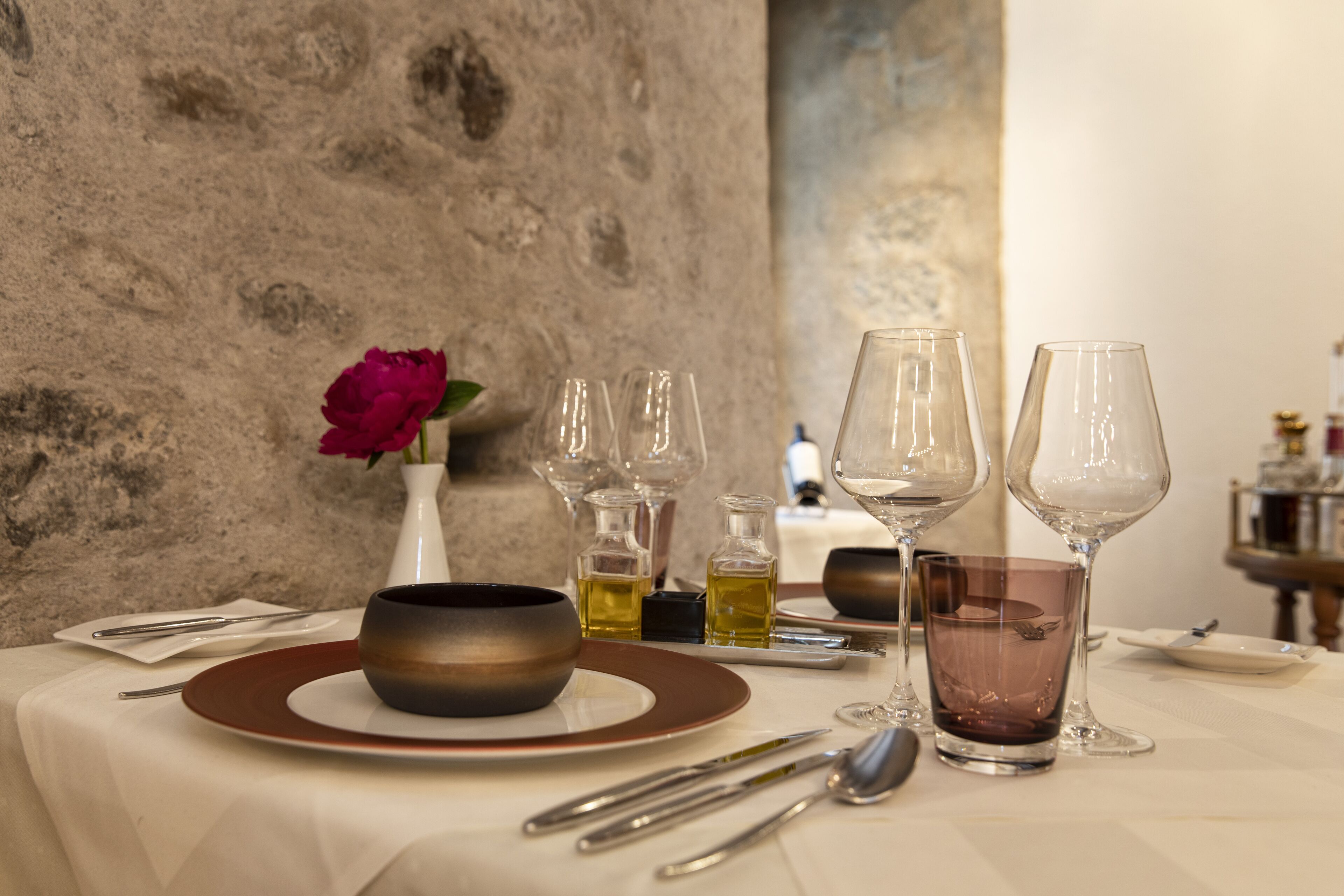 A table set with a bowl, plates, cutlery, and glasses, with a vase of flowers and bottles of oil on the table.