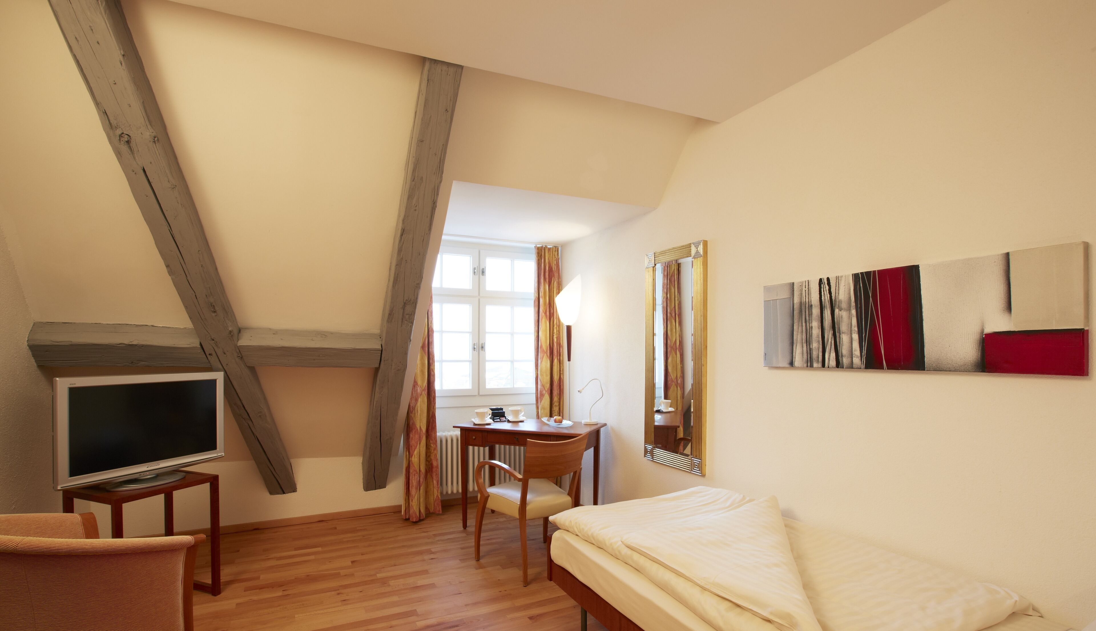 A bedroom with a wooden ceiling, a bed, a desk with a chair, a television, and a painting on the wall.