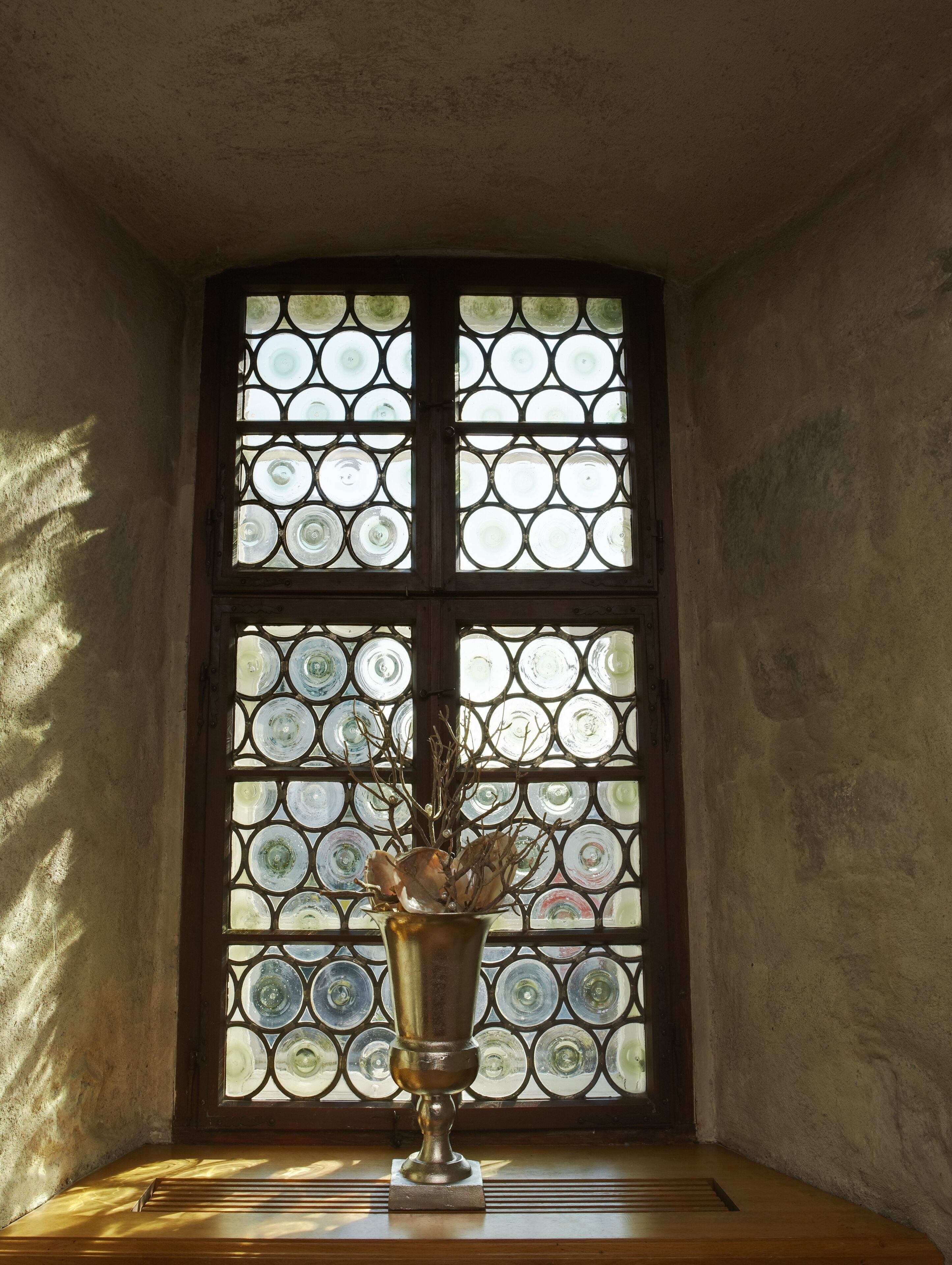A room with a large stained glass window, a metal vase with branches, and a wooden bench.