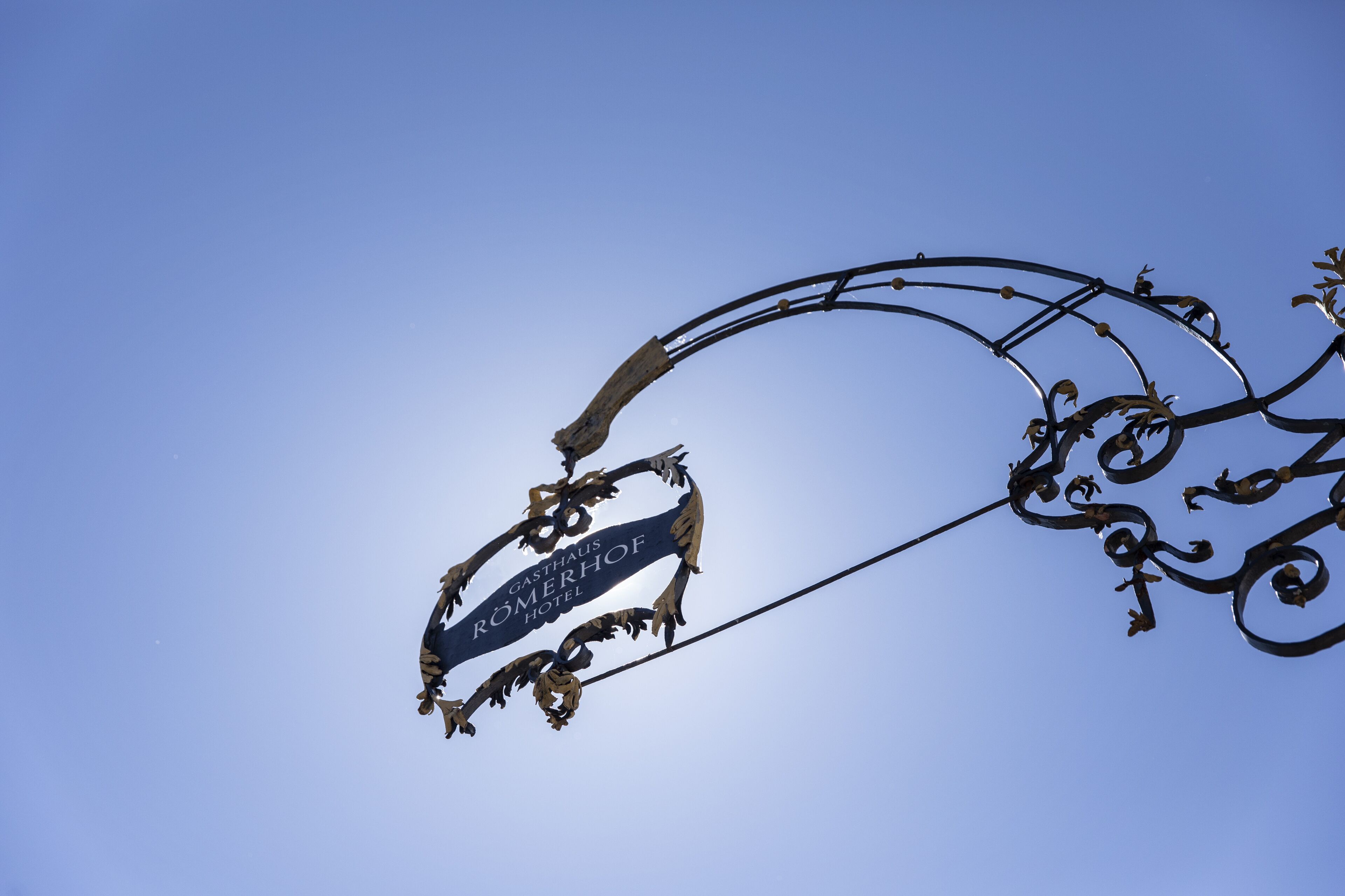 A wrought iron sign with the text "Gasthof Römerhof Hotel" against a clear blue sky.