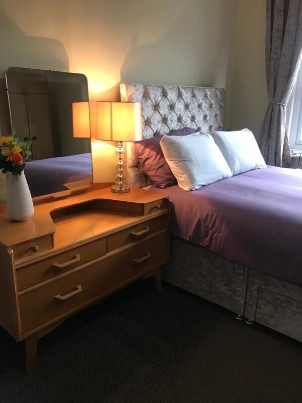 Una habitación de hotel con una cama, un aparador de madera con cajones, una mesita de noche con lámpara y un espejo.