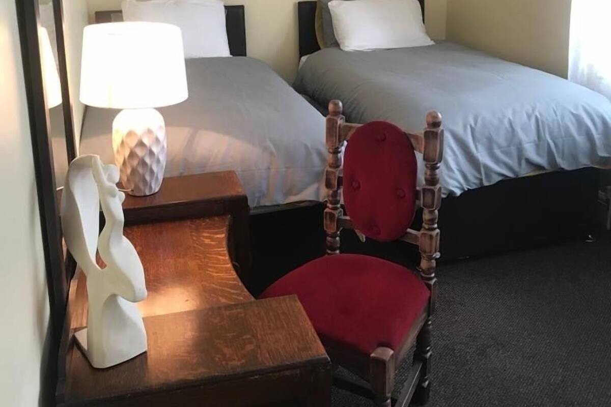 A hotel room with two beds, a wooden chair with a red cushion, a small table with a lamp, and a framed picture on the wall.