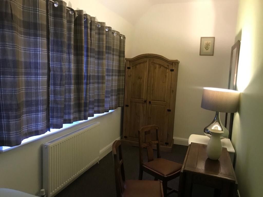 Ett rum med ett fönster med rutiga gardiner, ett element, en trägarderob, en stol, ett sängbord med en lampa och en inramad tavla på väggen.
