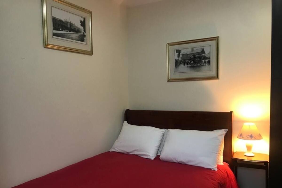A bed with a red bedspread and white pillows, a nightstand with a lamp, and two framed pictures on the wall.