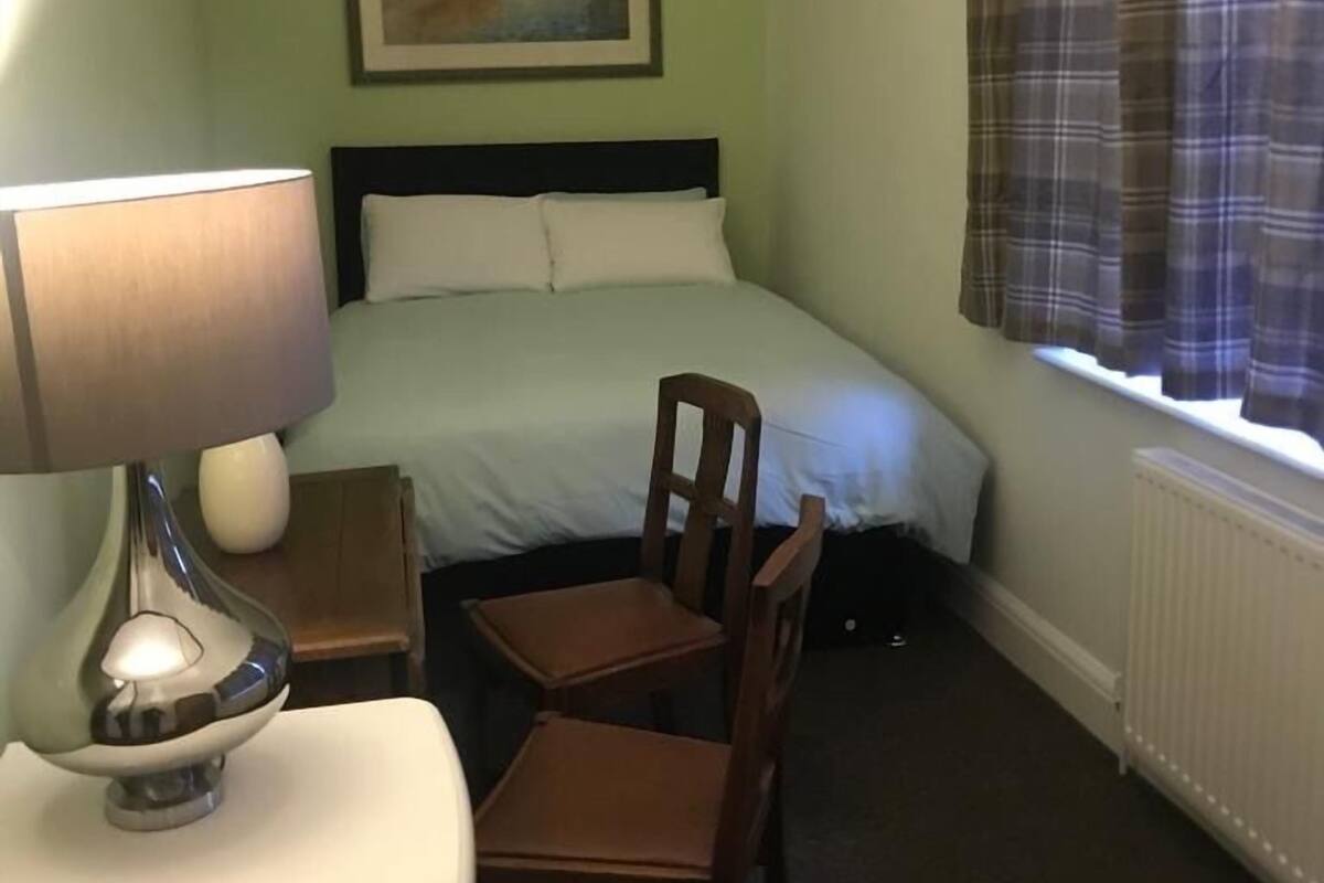 A hotel room with a bed, a desk with a lamp, a chair, a painting on the wall, and a window with curtains.