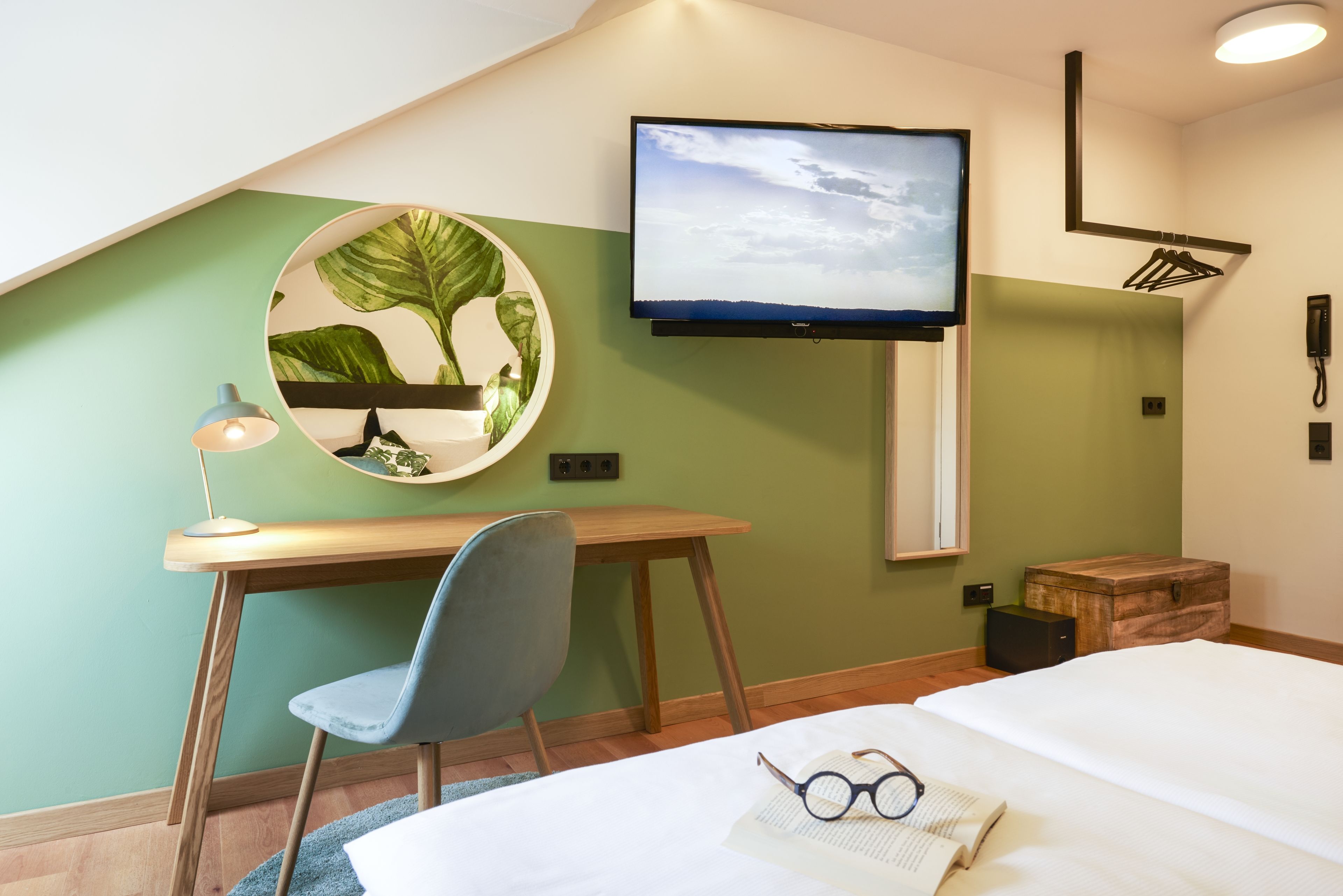 A hotel room with a bed, a desk with a chair, a television mounted on the wall, and a small wooden chest.