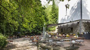 An outdoor seating area with tables and chairs, surrounded by trees and a stone building. - QUARTIER 82 by Arcadeon (Hagen)