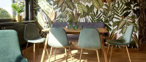 A dining area with a wooden table, four chairs, and a bench. The room features a large window, a radiator, and a potted plant.