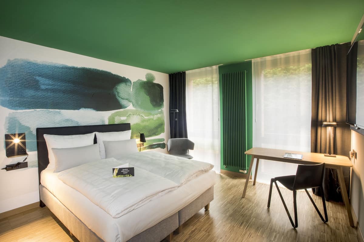 Une chambre d’hôtel avec un lit, un bureau, une chaise, une table de chevet et un tableau au mur.