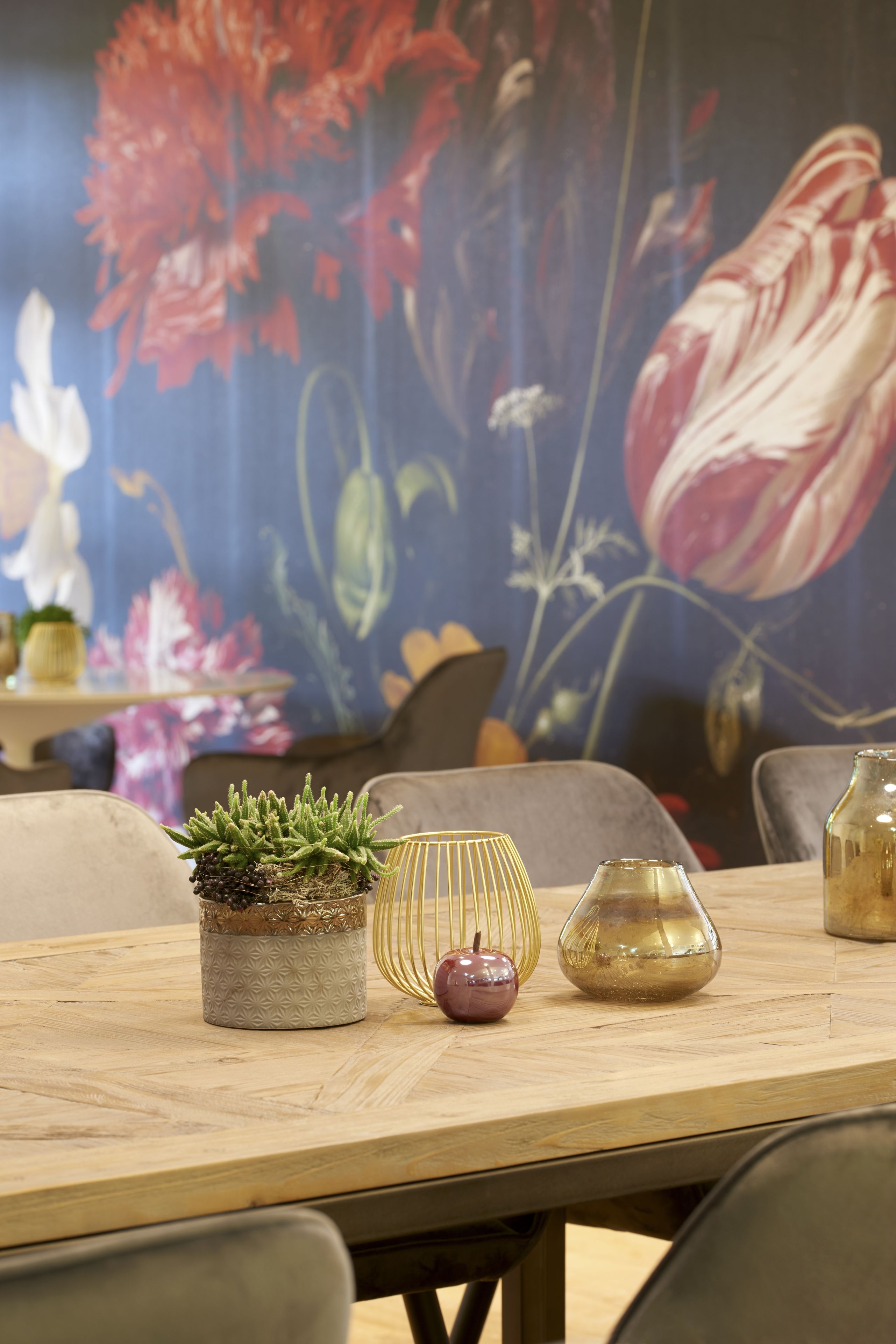 A dining table with a potted plant, a vase, and a candle. A floral mural is in the background.