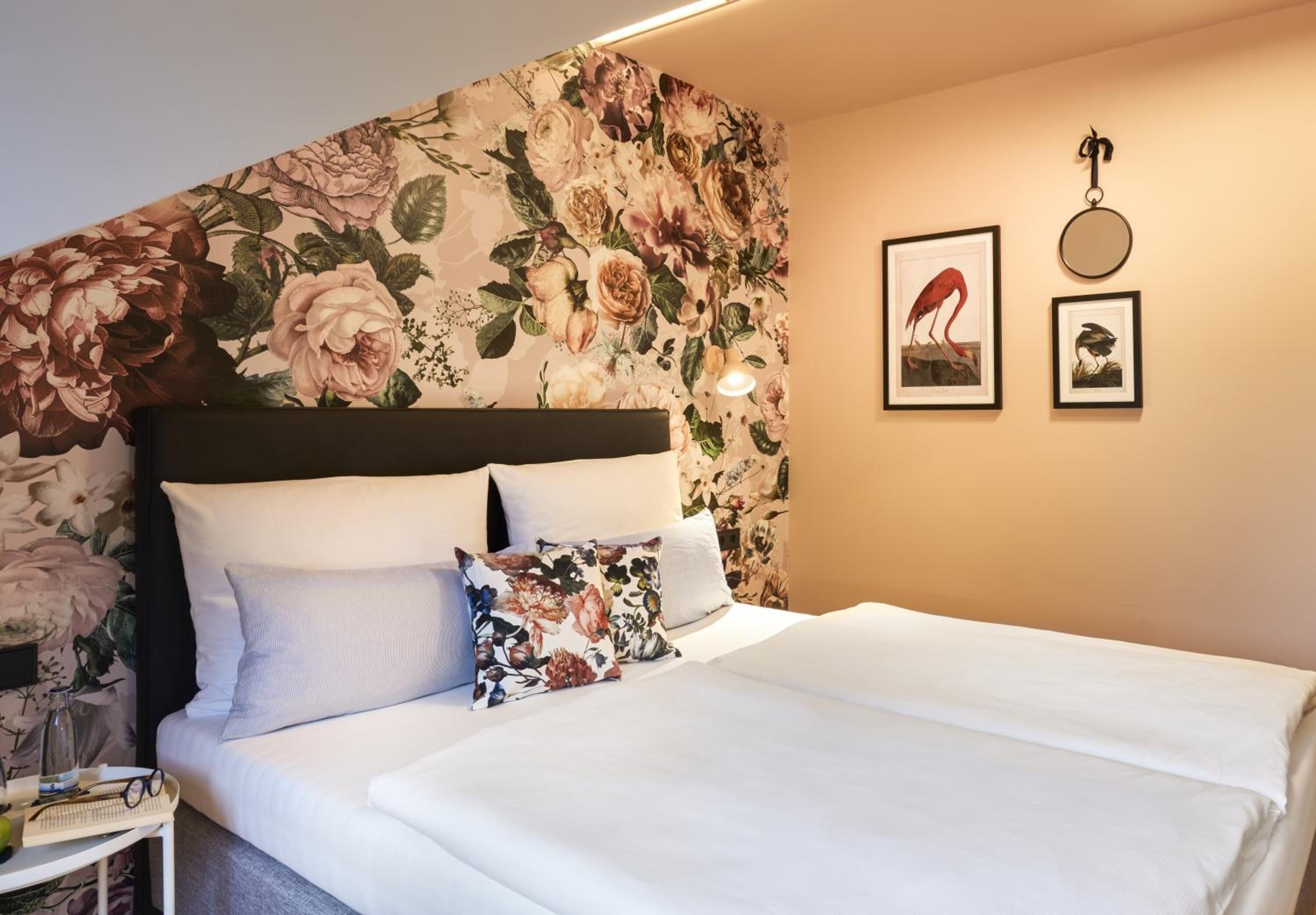 Un lit bien fait, avec du linge de lit blanc et des coussins à motifs floraux, une table de chevet avec une lampe, et un mur recouvert d’un papier peint floral et orné de tableaux encadrés.