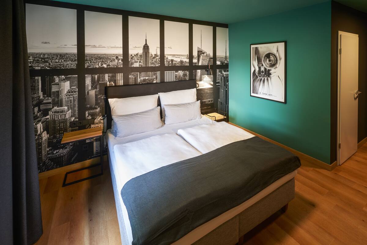 Un lit bien fait, avec une fresque représentant un paysage urbain sur le mur derrière, une œuvre d’art encadrée sur le mur et une porte sur la droite.