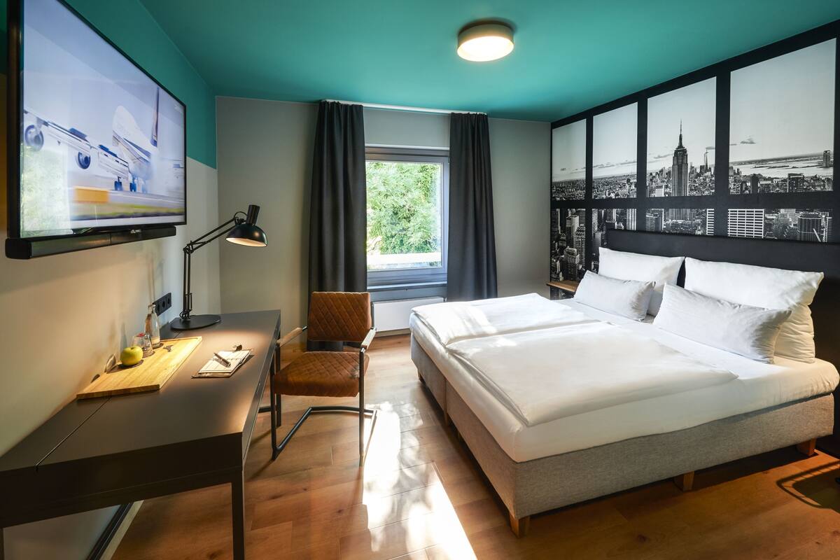 Une chambre d’hôtel équipée d’un lit, d’un bureau, d’une chaise, d’une télévision et ornée d’une fresque représentant un paysage urbain.