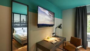 A hotel room with a bed, a desk with a lamp, a television, and a view of the cityscape. - QUARTIER 82 by Arcadeon (Hagen)