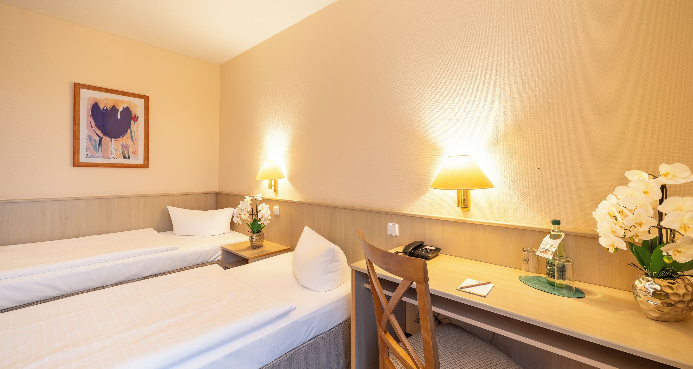 Habitación de hotel con dos camas, un escritorio, una silla, una lámpara, un teléfono, un florero con flores y un cuadro en la pared.