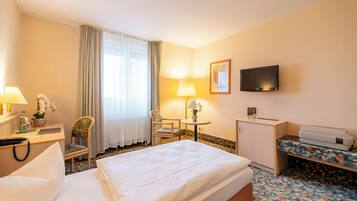 A hotel room with a bed, a desk with a lamp, a chair, a television, and a small table with a lamp and flowers.