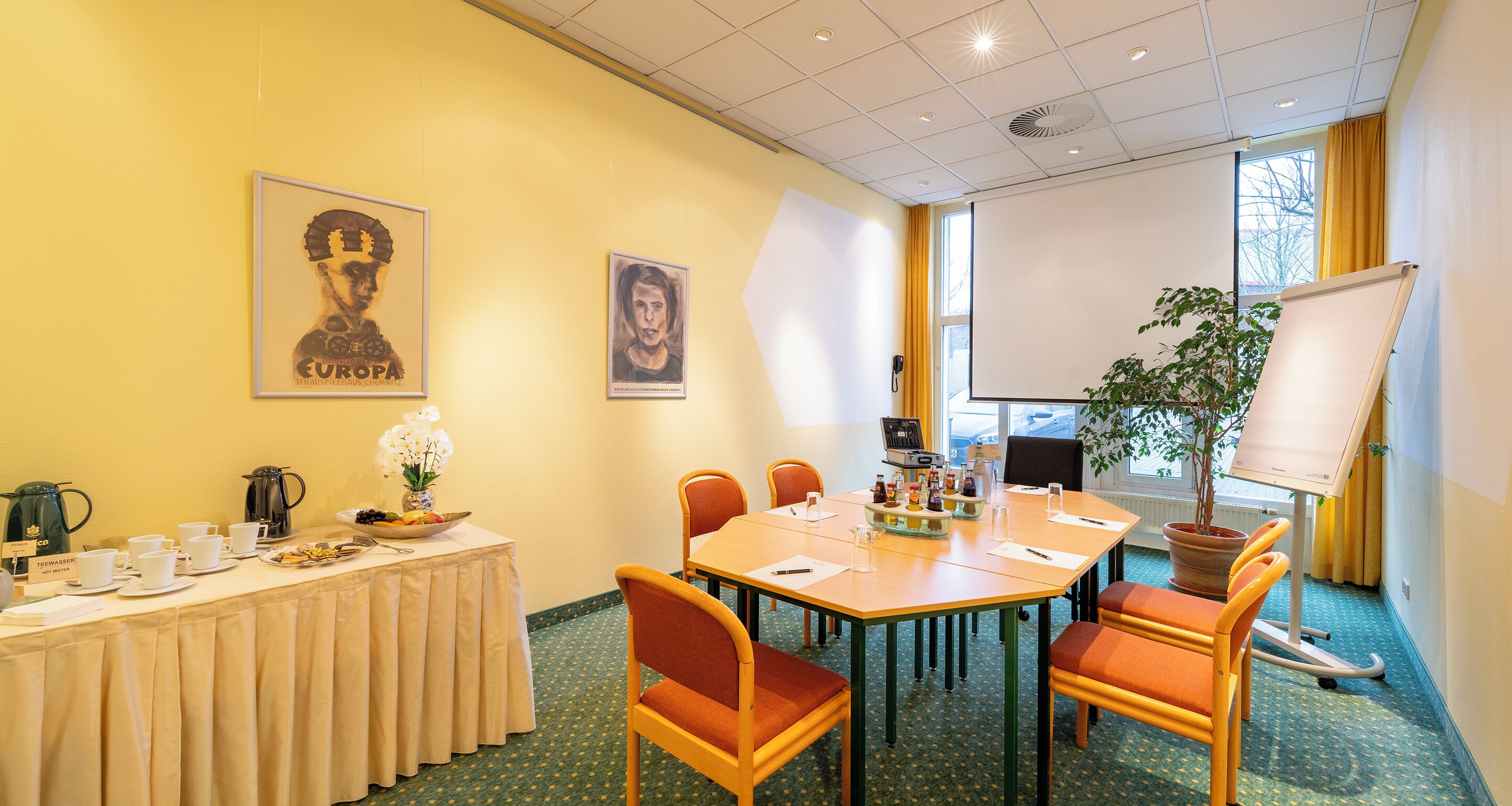 A conference room with a table set for a meeting, chairs, a projector screen, and a plant.