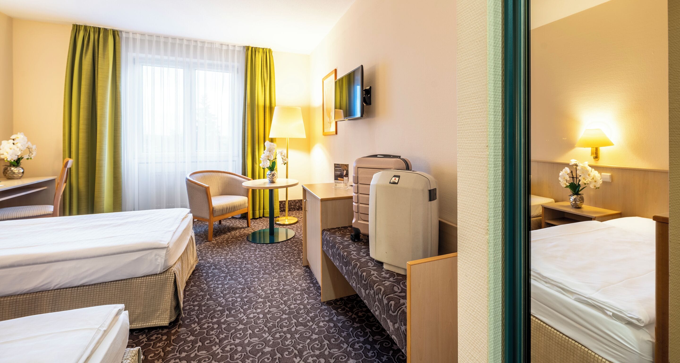 A hotel room with two beds, a desk, a chair, a small table, a lamp, a TV, and a window with curtains.