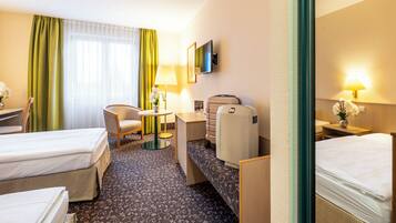 Ein Hotelzimmer mit zwei Betten, einem Schreibtisch, einem Sessel, einem kleinen Tisch, einer Lampe, einem Fernseher und einem Fenster mit Vorhängen.