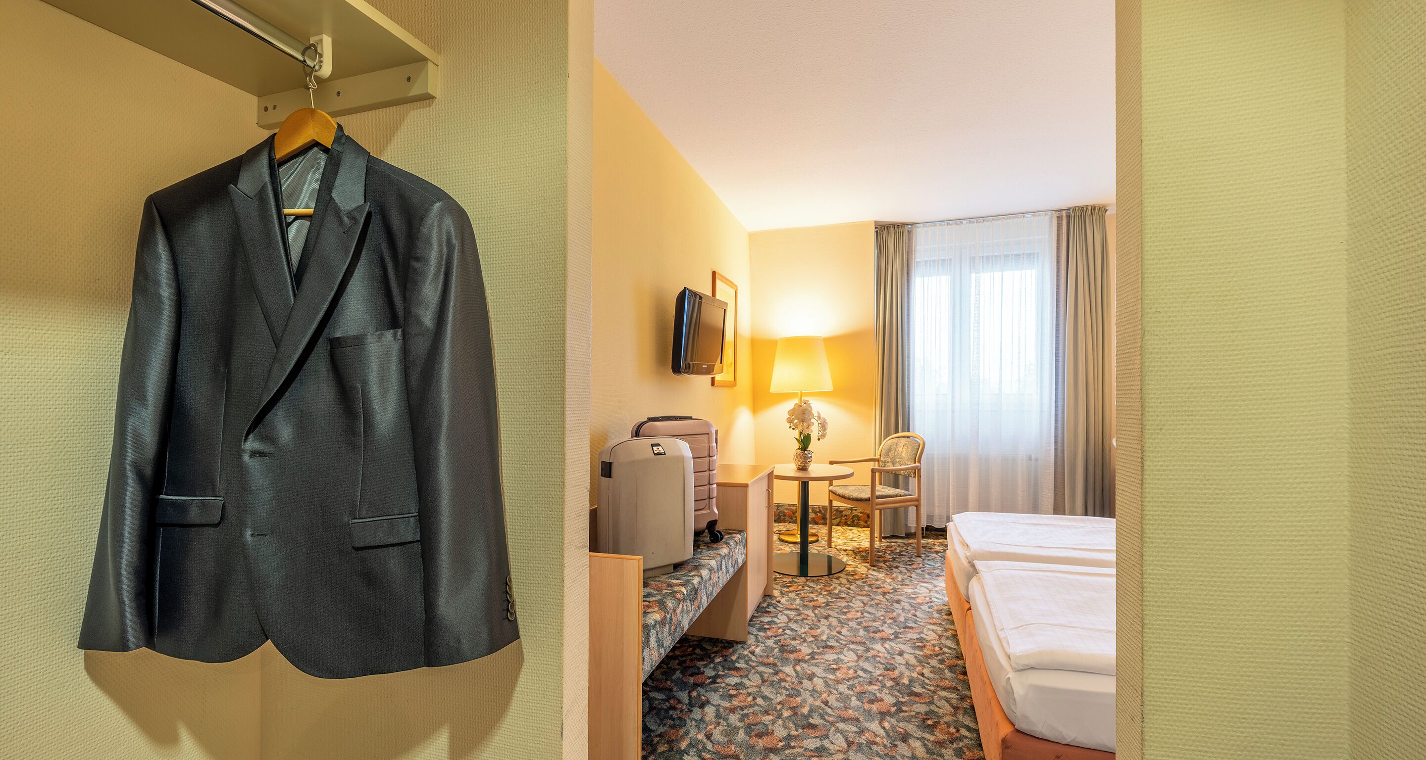 Ein Hotelzimmer mit einem Bett, einem Schreibtisch mit Lampe, einem Sessel, einem Koffer und einem Mantel, der an der Wand hängt.