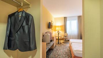 Ein Hotelzimmer mit einem Bett, einem Schreibtisch mit Lampe, einem Sessel, einem Koffer und einem Mantel, der an der Wand hängt.
