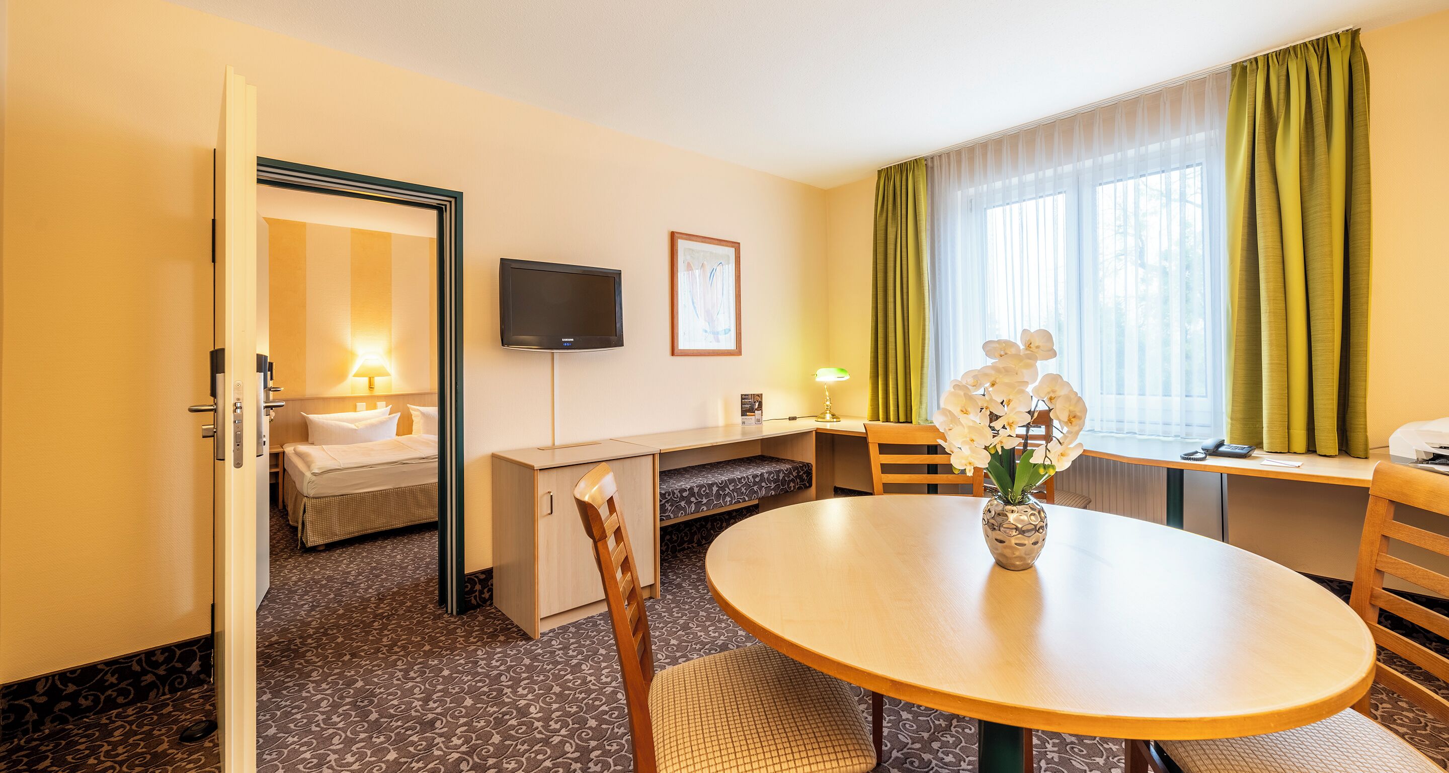 A hotel room with a bed, a desk, a television, a vase with flowers, and a window with curtains.