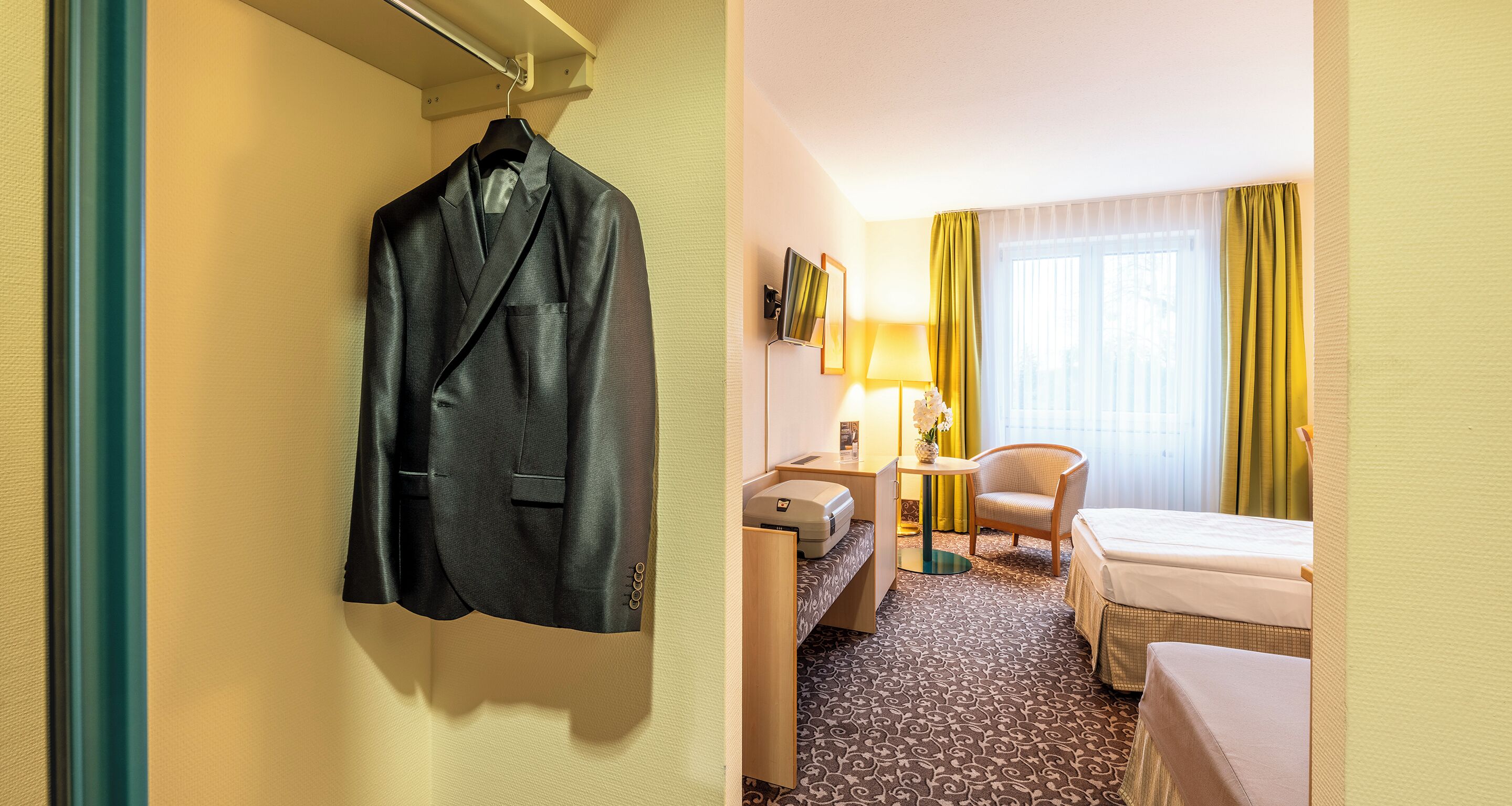 Une chambre d’hôtel avec un lit, un bureau, une chaise, une lampe et un manteau accroché au mur.