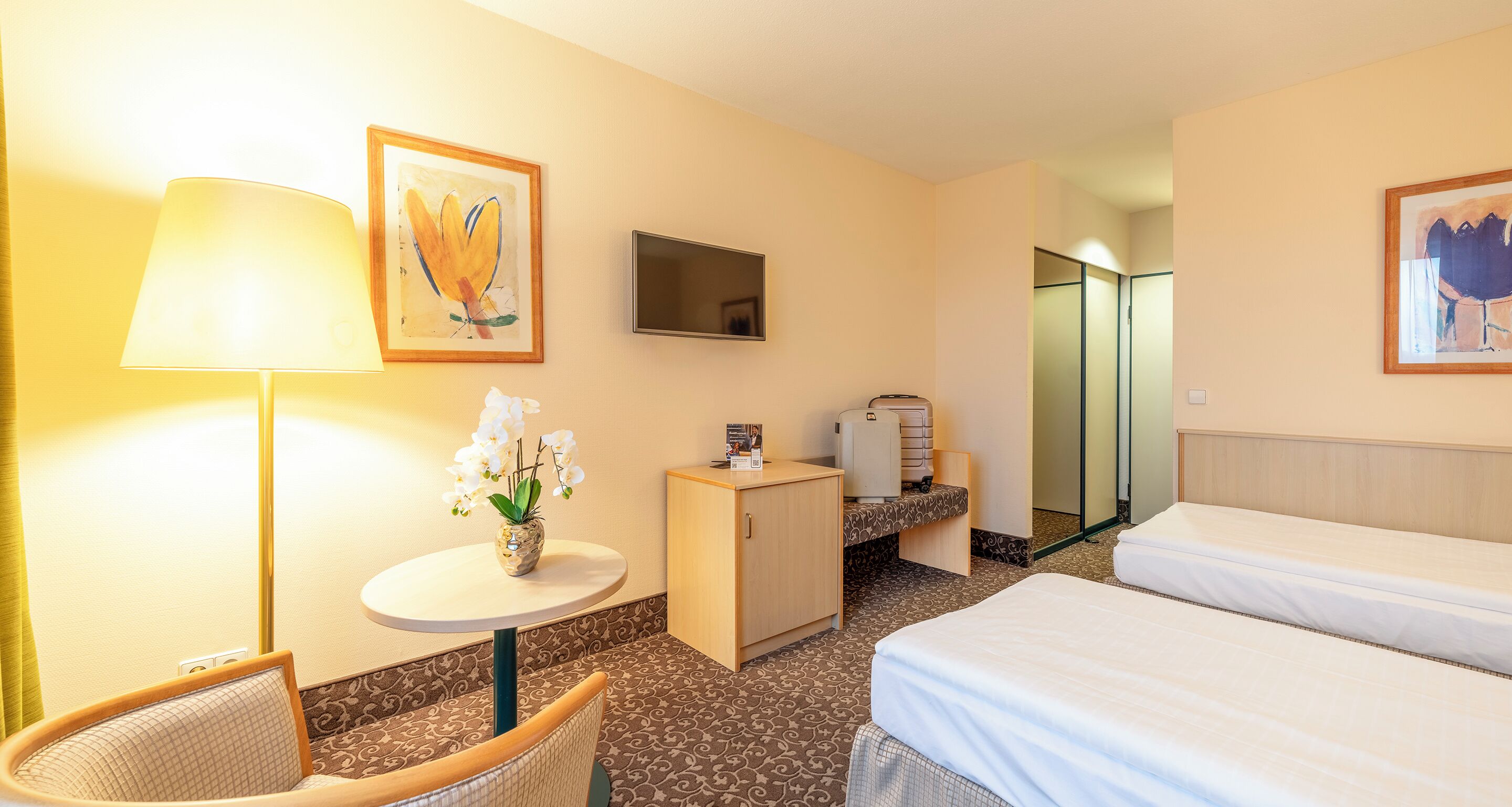 A hotel room with a bed, a small table with a lamp, a cabinet, and a mirror.