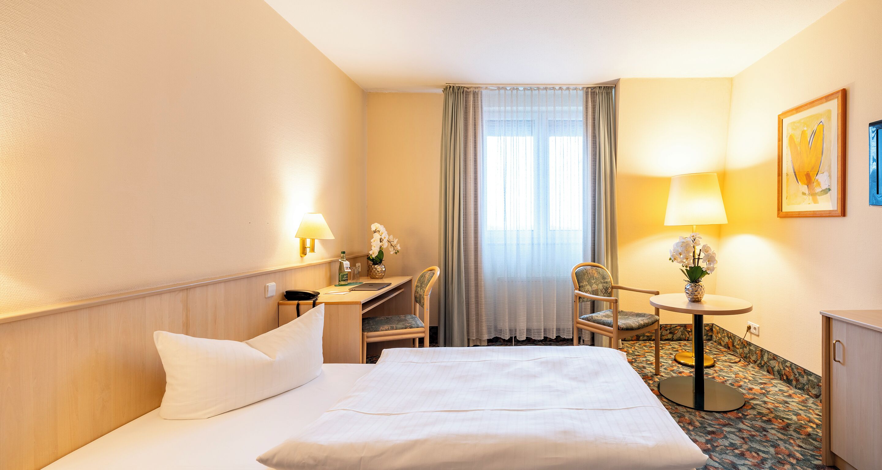Une chambre d’hôtel comprenant un lit, un bureau, une chaise, une petite table avec une lampe et une fenêtre avec des rideaux.