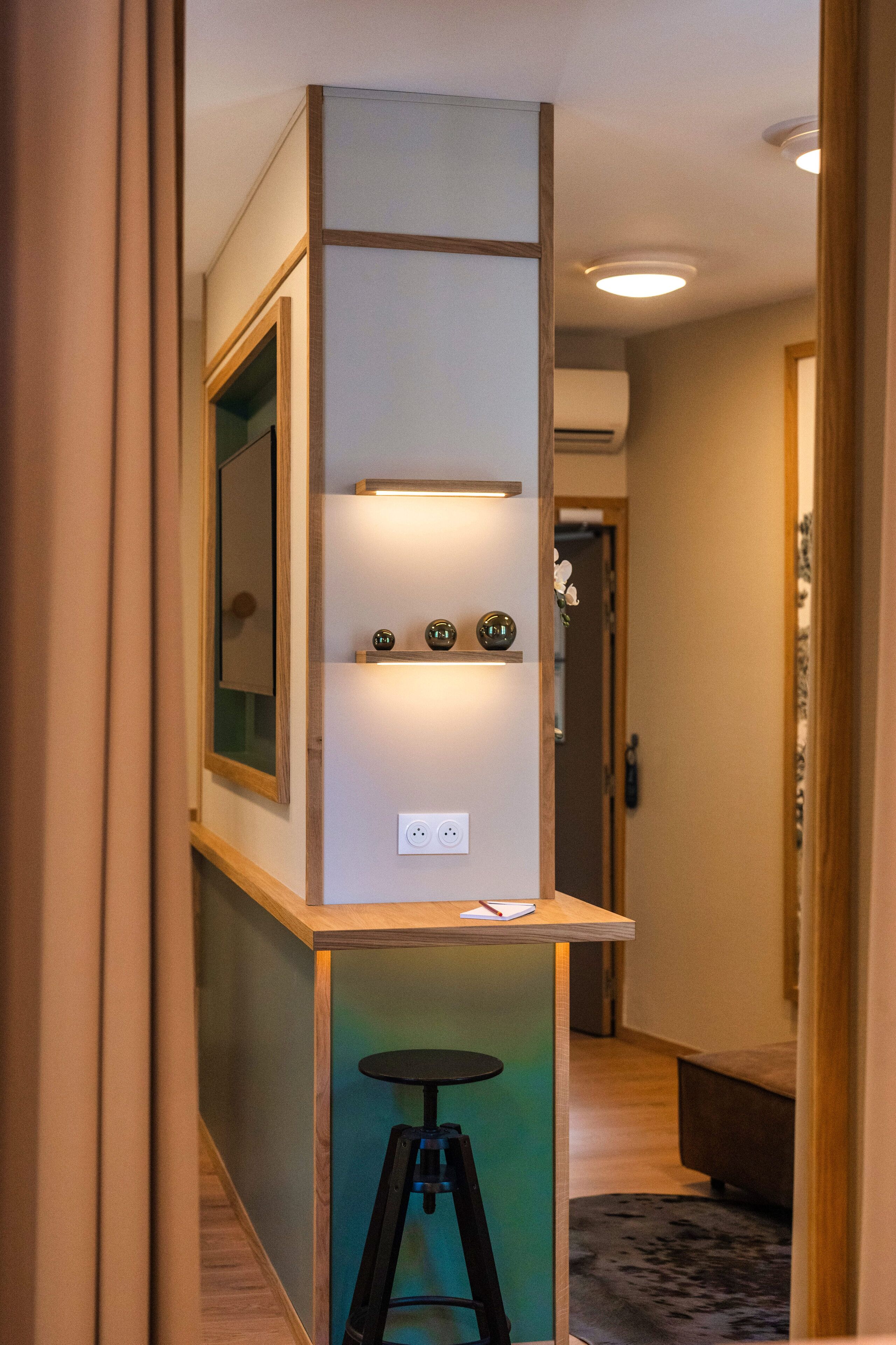 A narrow hallway with a small bar area featuring a stool, a shelf with decorative items, and a mirror.