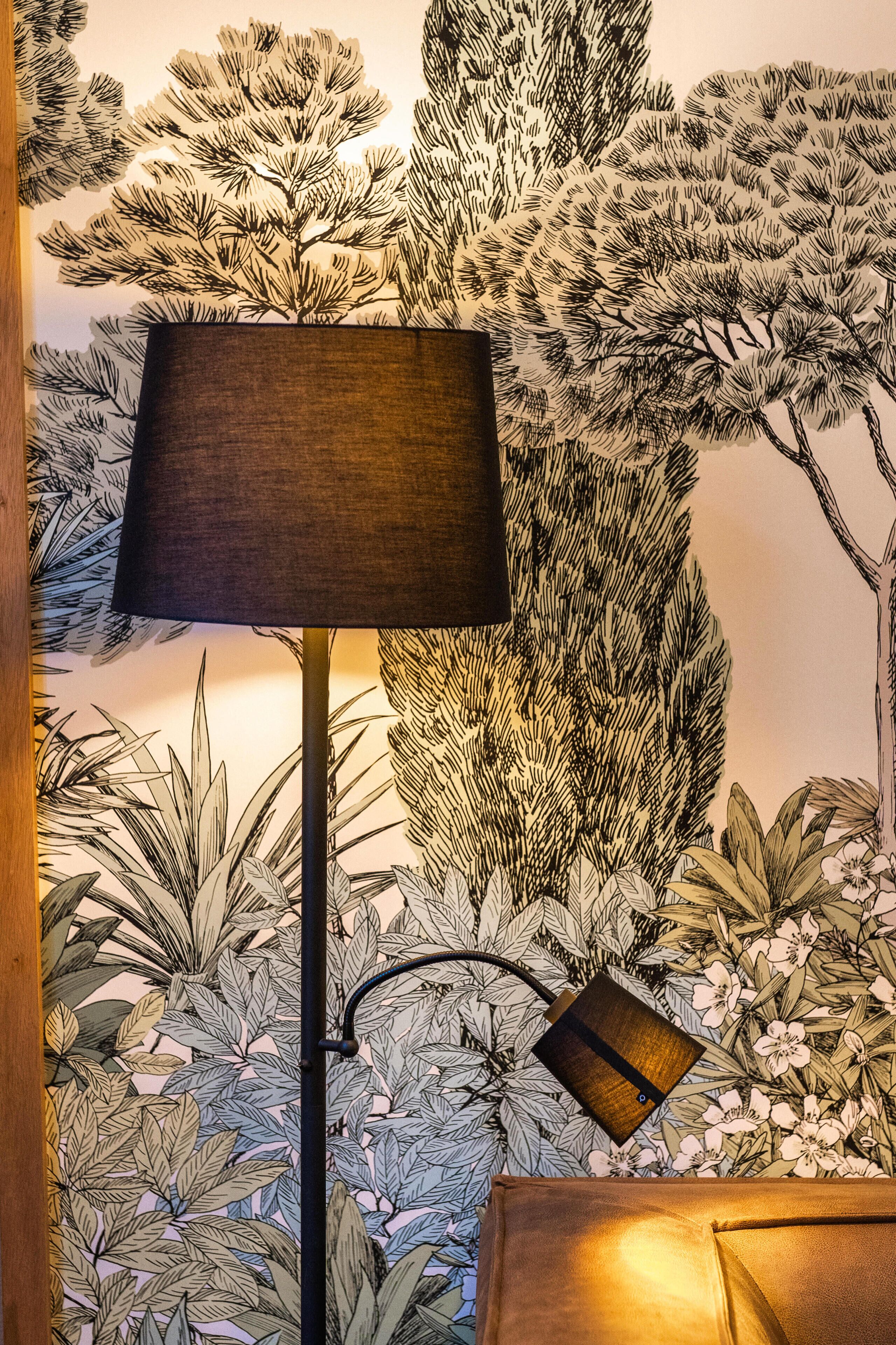 A lamp with a dark shade on a wall with a botanical wallpaper.