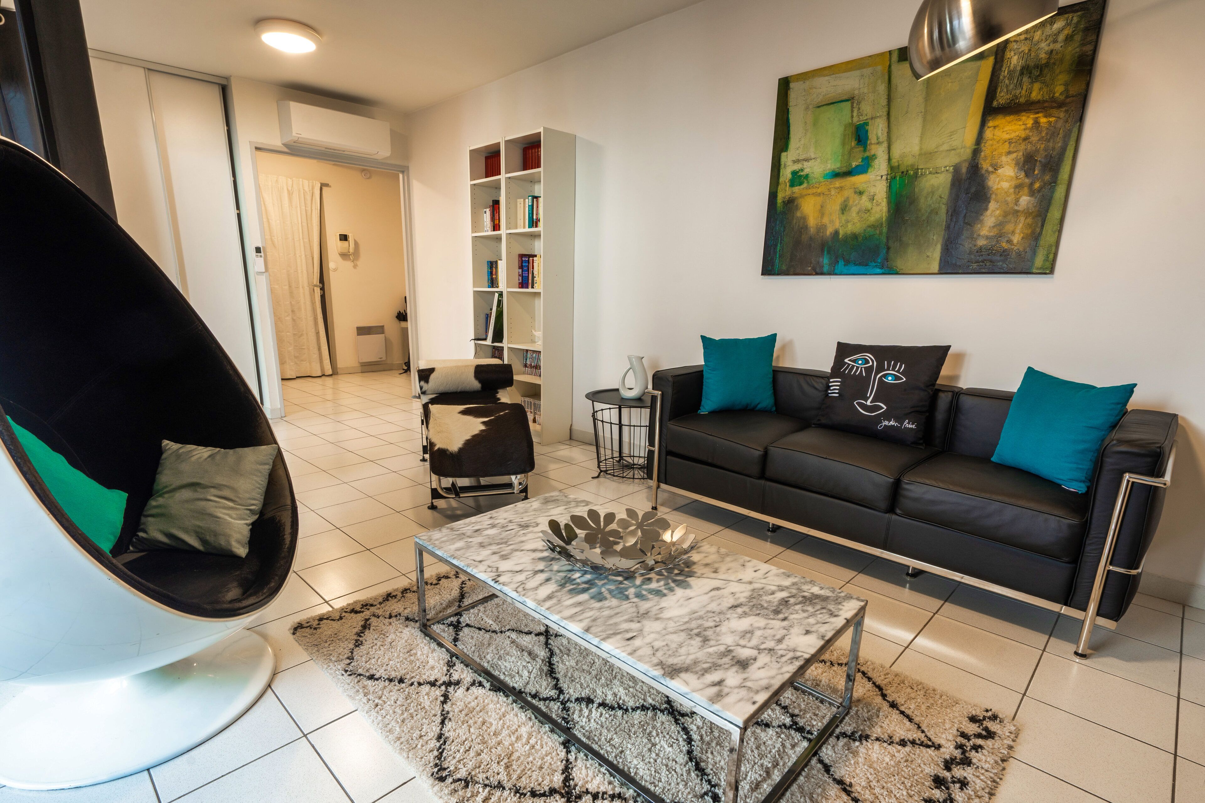 A modern living room with a black leather sofa, a white chair, a glass coffee table, and a bookshelf.