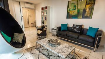 A modern living room with a black leather sofa, a white chair, a glass coffee table, and a bookshelf.