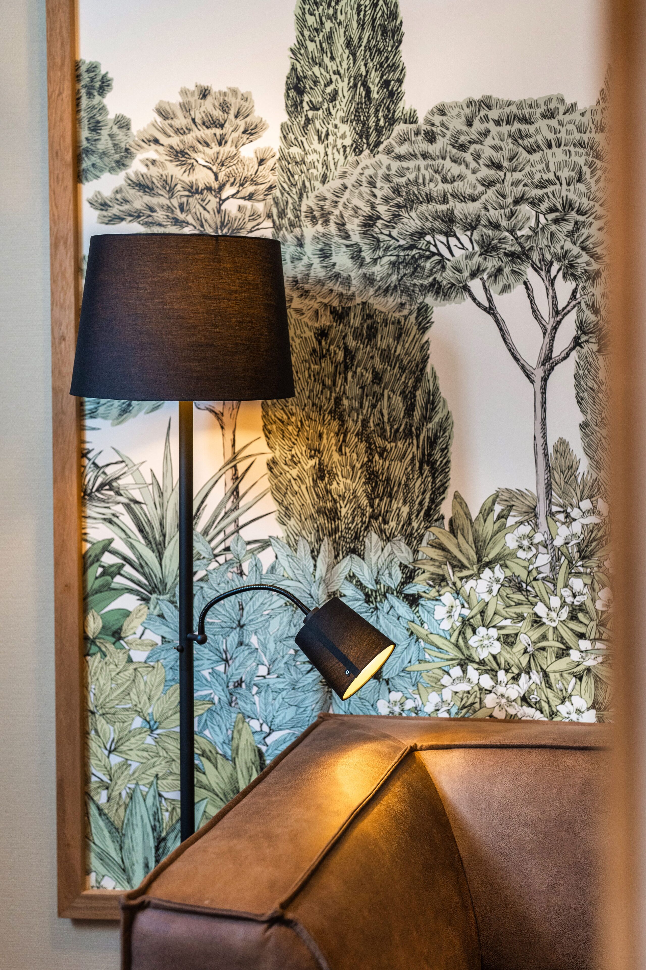Une lampe avec un abat-jour noir, posée sur une table en bois, devant un mur orné d’une fresque détaillée représentant des arbres et du feuillage.