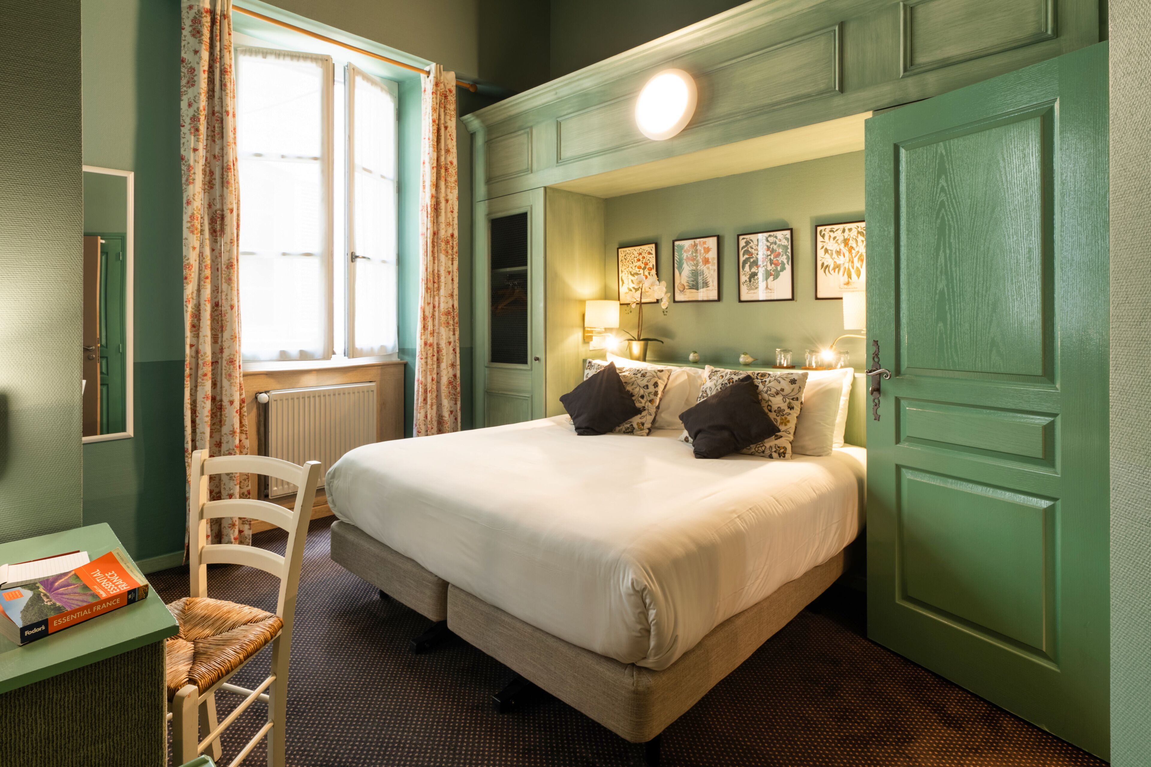 A hotel room with a large bed, a chair, a table with a book, and a window with curtains.