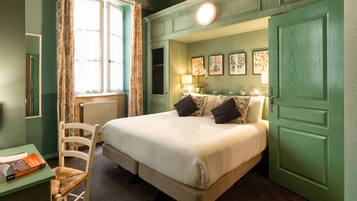 A hotel room with a large bed, a chair, a table with a book, and a window with curtains.
